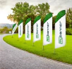 Flera beachflaggor med tryck från Mybanner uppställda utomhus på en grön gräsmatta. Flaggorna har en vit bakgrund med gröna detaljer och företagslogotyp. Perfekt för event, mässor och utomhusreklam.